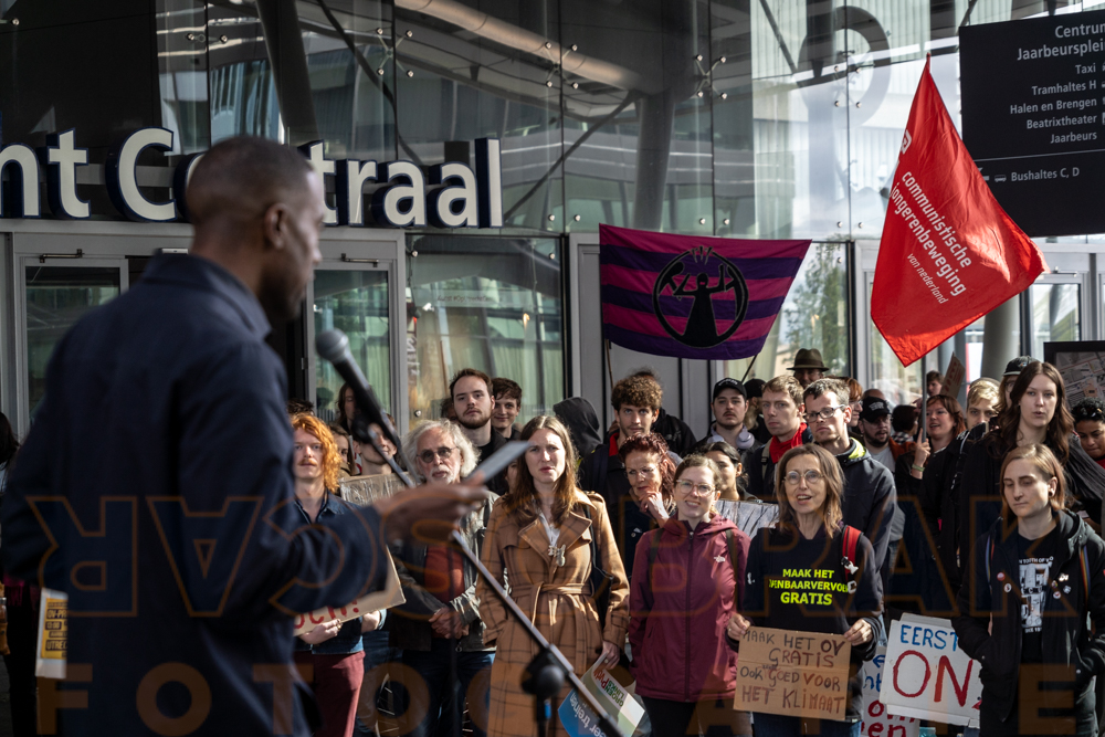 Foto van een spreker en de aanwezigen van de OV toegankelijkheidsdemo. Op de achtergrond is de mad pride vlag zichtbaar.