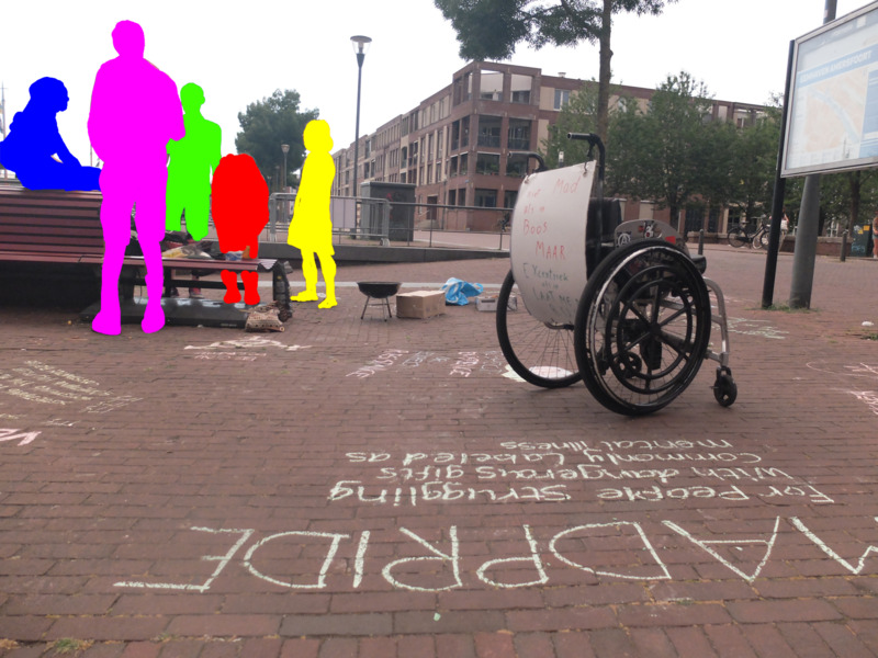 Vijf mensen staan/zitten rond een bankje te praten met naast hen een barbeque. Op de voorgrond staat een rolstoel met protestbord. Op de grond zijn met krijt teksten geschreven. Een daarvan leest "madpride".