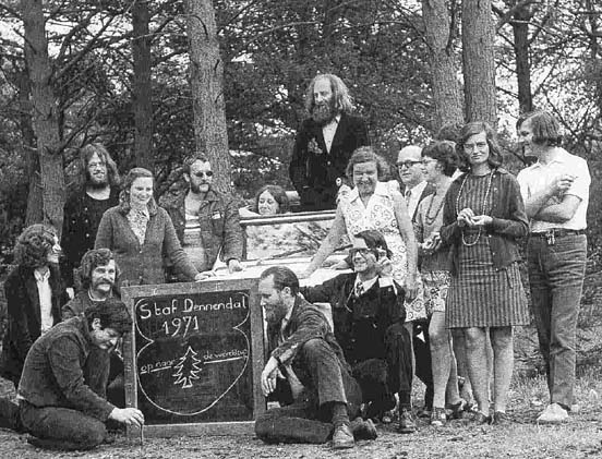 Zwart-wit foto van mensen die staan of zitten op het gras, met achter hen dennenomen. Tussen hen in staat een krijtbord met daarop "Staf Dennendal 1971".
